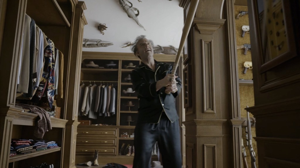 A man in black pajamas stands in a wooden closet, holding a baseball bat, with shelves of clothing and displayed weapons in the background.